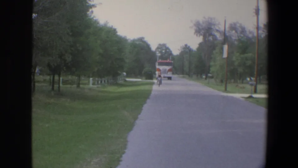 1973 Minnesota home-movie frame showing a cyclist on a quiet road with trees, lawns, utility poles, and a distant fire truck.