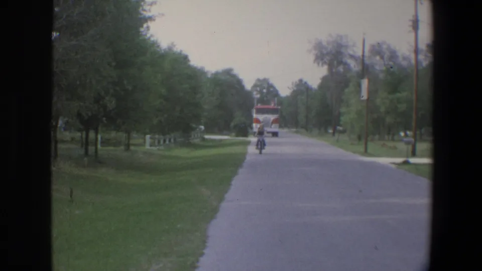 1973 Minnesota 8mm frame of a cyclist riding along a tree-lined street, with a red-and-white fire truck visible in the background.