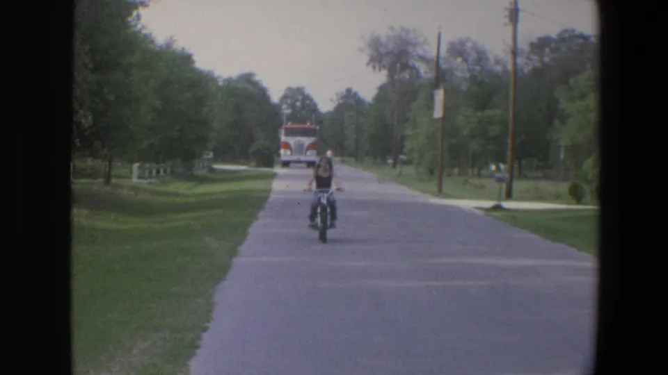 1973 Minnesota 8mm frame showing a motorcycle rider near the camera on a quiet street, with lawns, trees, utility poles, and a distant fire truck.