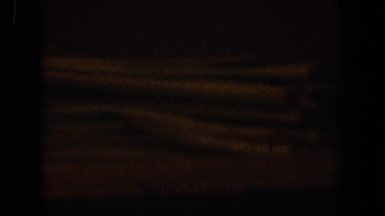 Dark 1958 Lubbock, Texas 8mm frame with faint orange light, indistinct horizontal forms, and heavy shadow across the view.