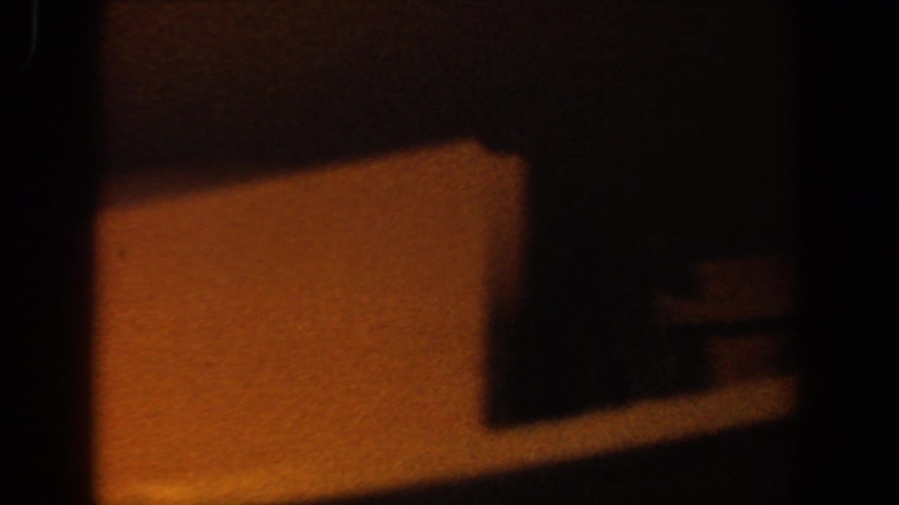 1958 Lubbock 8mm frame showing a dim interior corner with a white shelf or ledge, amber light, and deep surrounding shadow.
