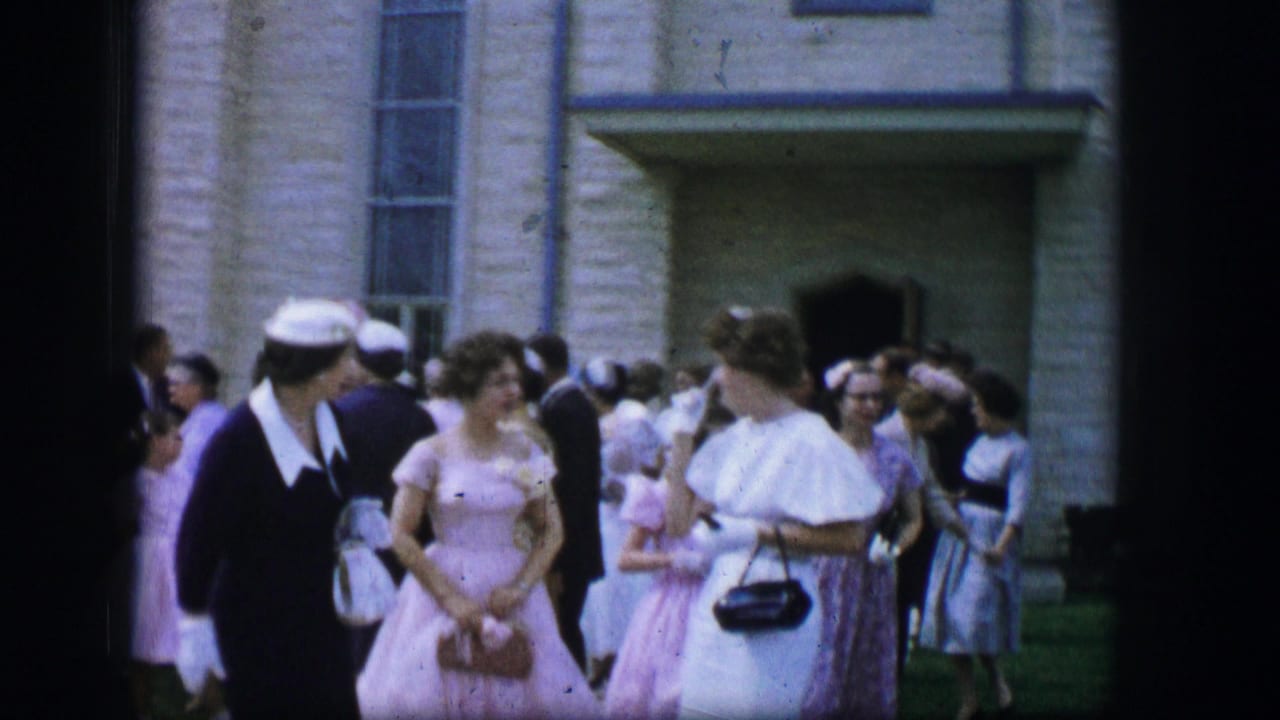 Women in formal dresses and hats stand outside a stone church entrance with other attendees nearby.
