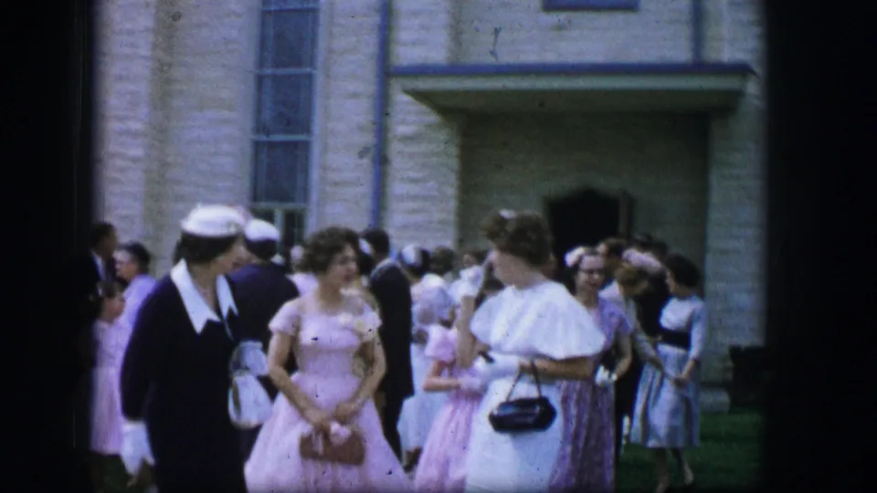 1962 Moberly, Missouri home-movie frame showing women in pastel formal dresses and hats talking outside a stone church.