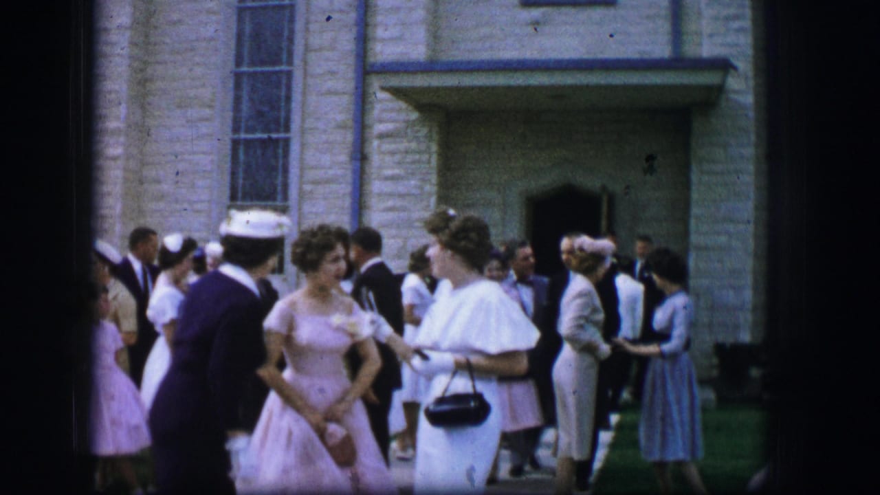 Two women in light formal dresses stand near a church doorway as a crowd gathers around them.