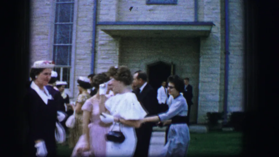 1962 Moberly, Missouri 8mm frame of wedding guests in hats, gloves, dresses, and suits chatting near a stone church doorway.