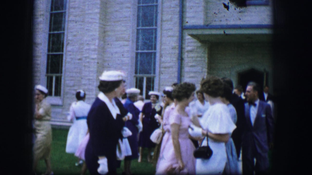 Formal wedding guests stand in small groups on the lawn outside a church.