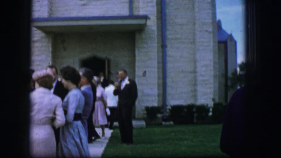 1962 Moberly, Missouri 8mm frame of wedding guests conversing near the church entrance, partly framed by dark film edges.