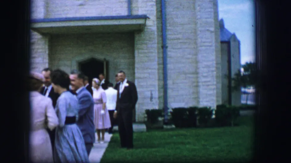 1962 Moberly, Missouri frame with wedding guests in suits, dresses, and hats standing on the lawn outside a stone church.