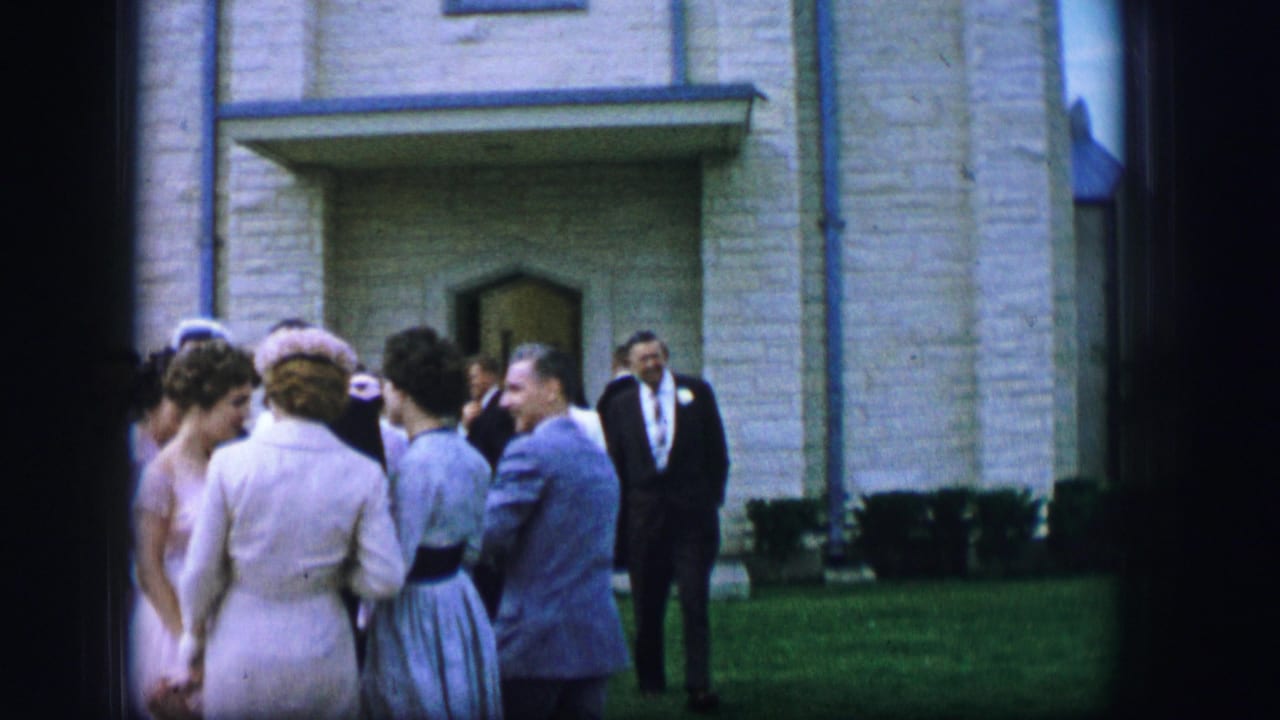 Adults in pastel dresses and suits talk outside a stone building.