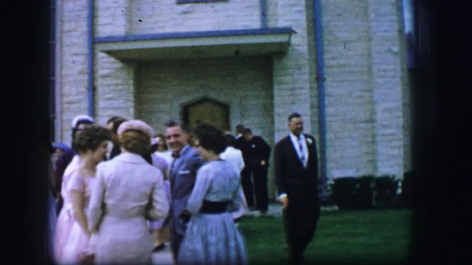 Adults in dresses and suits stand outside a pale stone building while a man in a dark suit walks through the frame.