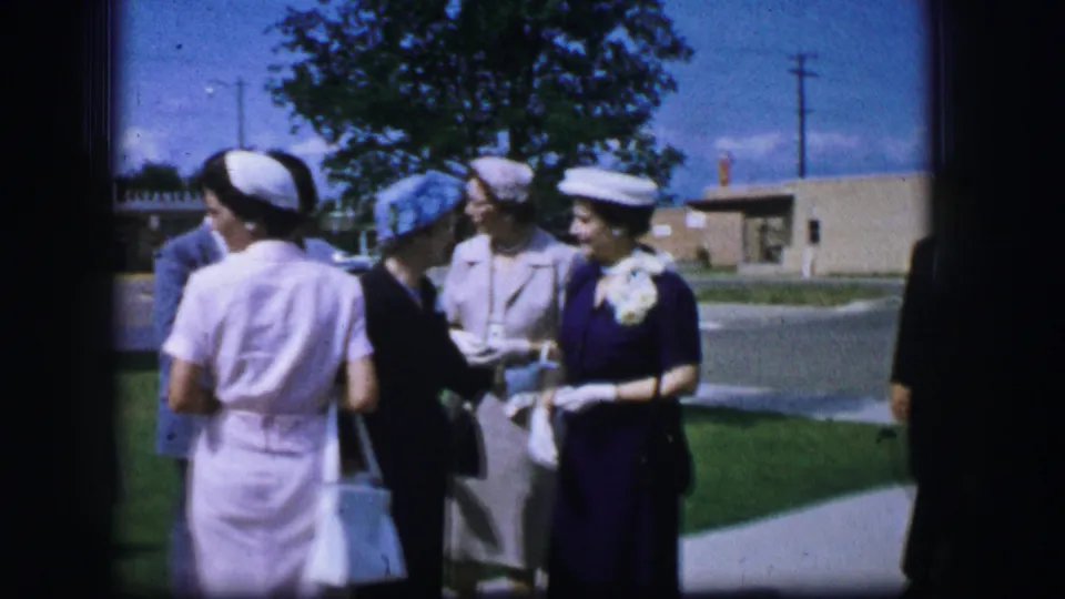 Formally dressed adults converse near a tree and road in Moberly, Missouri, with a woman in a white uniform seen from behind.