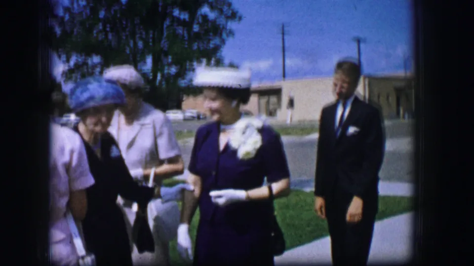 A woman in a dark dress with a white hat and large white flowers talks with others outdoors in 1962 Moberly, Missouri.
