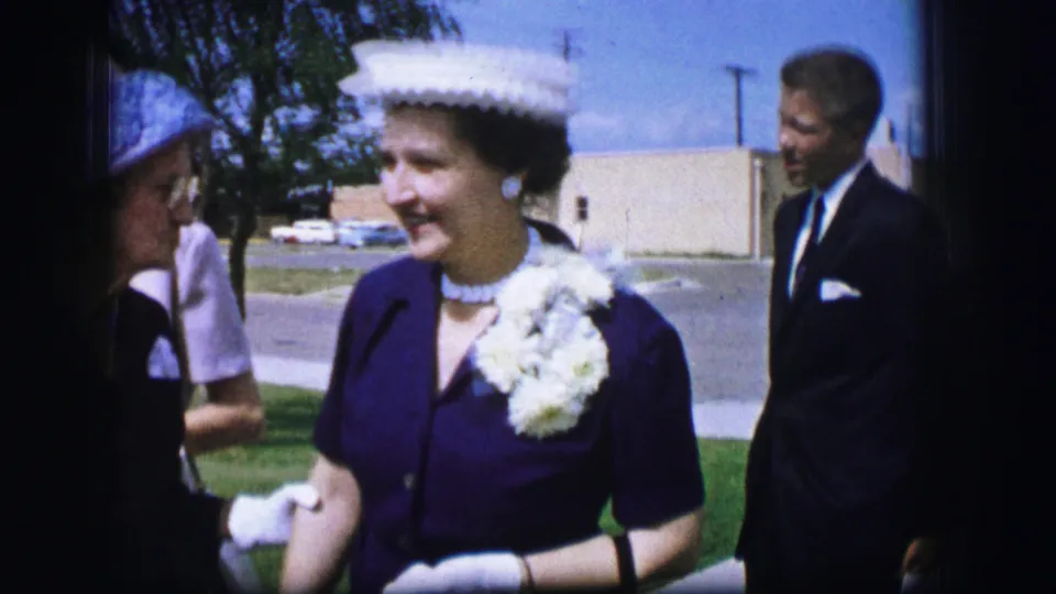 Close view of a smiling woman in a dark dress, white hat, gloves, and large white flowers in Moberly, Missouri, with a man in a suit behind her.