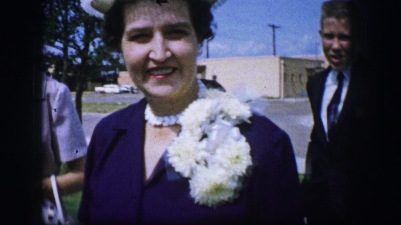In 1962 Moberly, Missouri, a woman in a dark purple dress and white corsage smiles close to the camera as guests stand behind her.