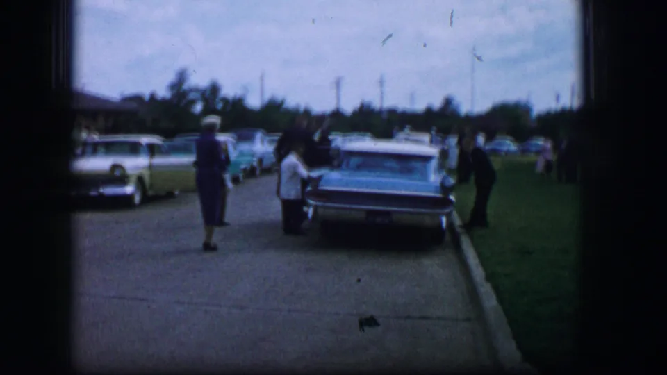 Wedding guests in 1962 Moberly, Missouri, stand and walk near parked mid-century cars on a quiet street beside grass.