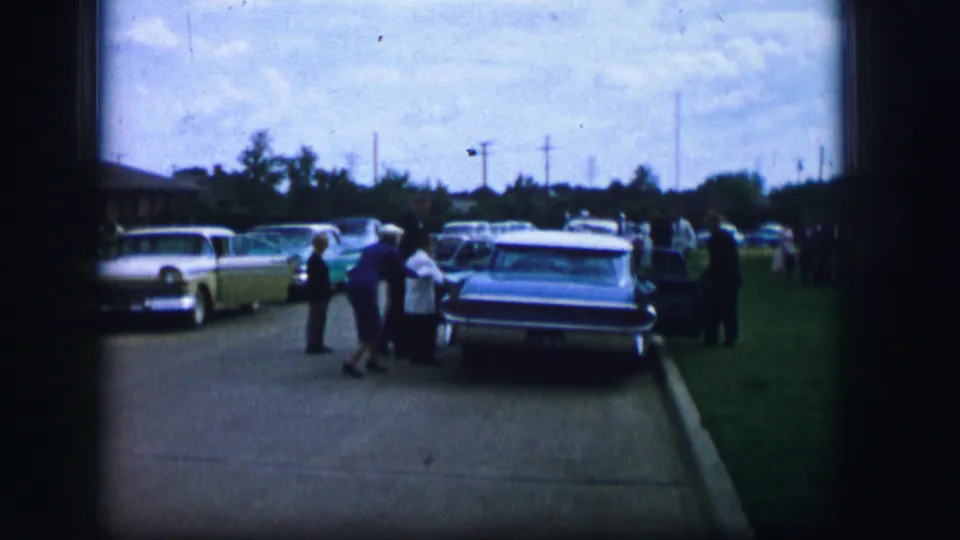 A small group of wedding guests stands by a parked car on a Moberly street, with additional 1960s cars visible along the curb.