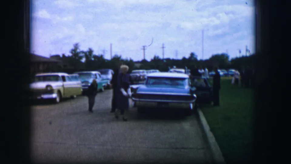 A woman in formal dress walks near the rear of a parked car as other wedding guests linger along the curb in Moberly, Missouri.