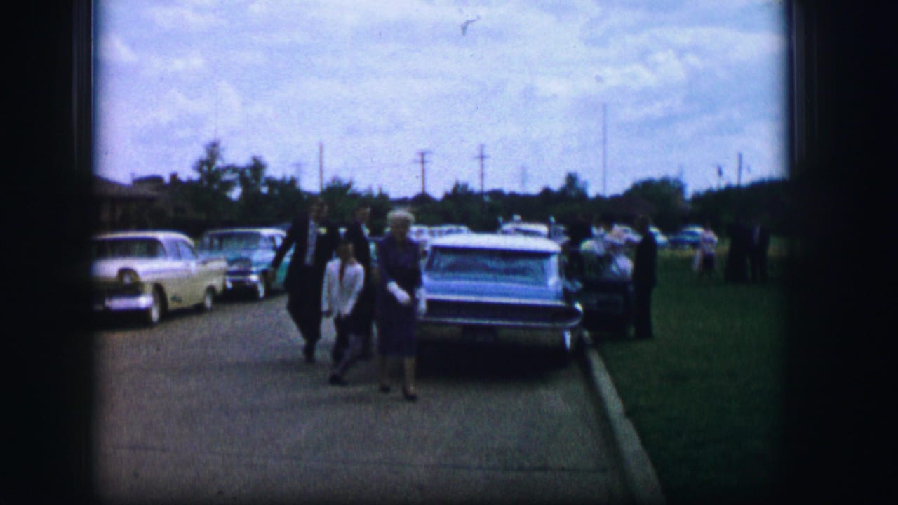 Woman in a blue dress walks on a sidewalk near parked mid-century cars and other pedestrians.