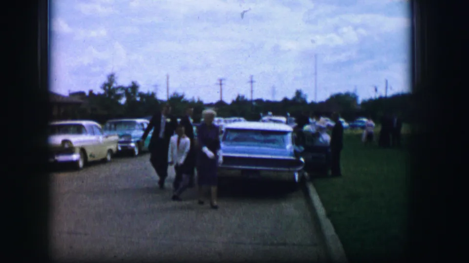 In Moberly, Missouri, in 1962, a woman in a blue dress walks near parked cars while adults move along the sidewalk and grass.