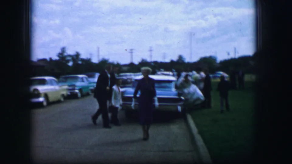 A woman in a blue dress approaches along the sidewalk in the 1962 Moberly footage, with suited pedestrians and parked cars behind her.