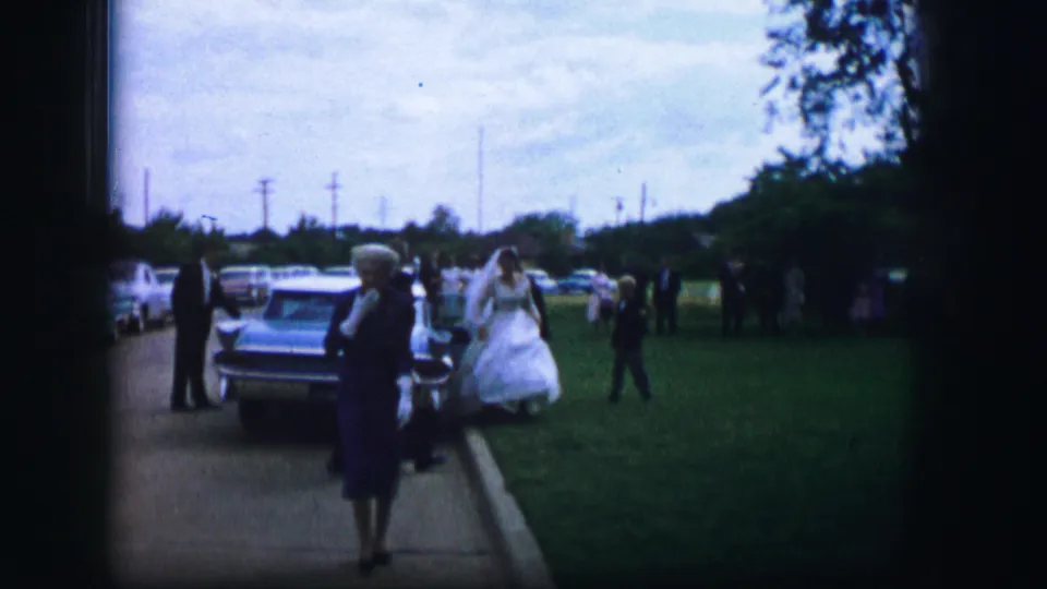 The woman in blue continues toward the camera in Moberly, Missouri, with a white-dressed figure and parked car visible behind her.