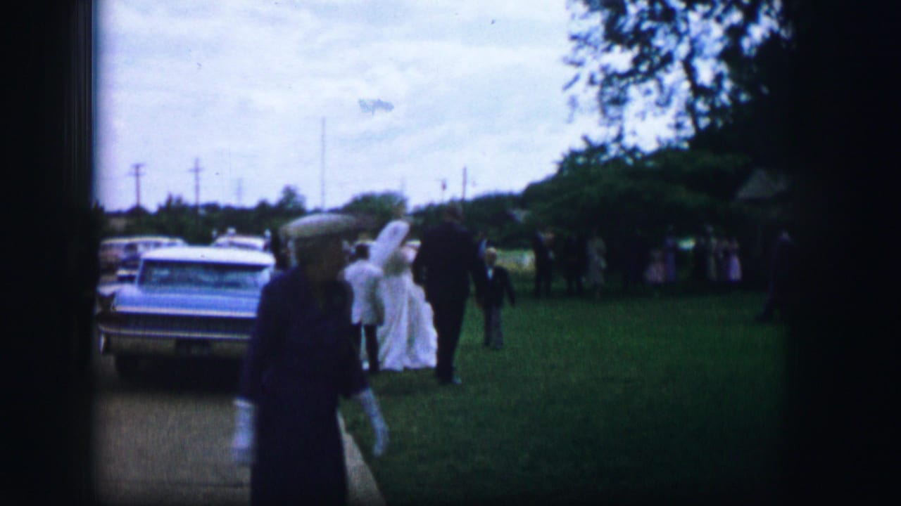In Moberly, Missouri, 1962, a bride in a white gown stands near a parked sedan while guests gather on a grassy wedding site.