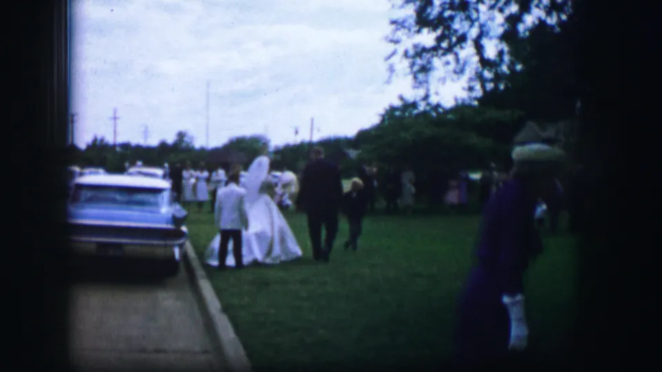 The bride and attendants move across a lawn near a parked 1960s sedan, with wedding guests visible in the background.