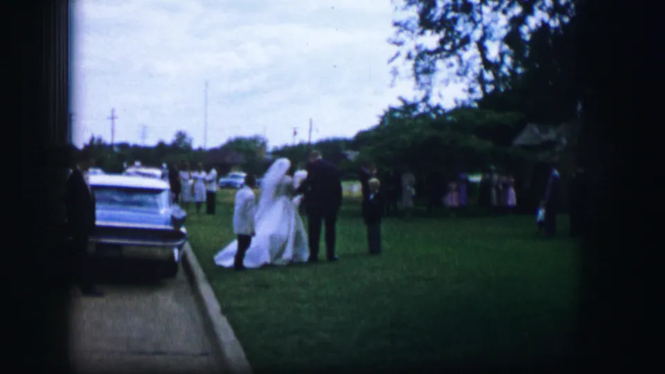 In the 1962 Moberly wedding footage, the bride stands beside a suited man near a sedan as guests look on from the lawn.
