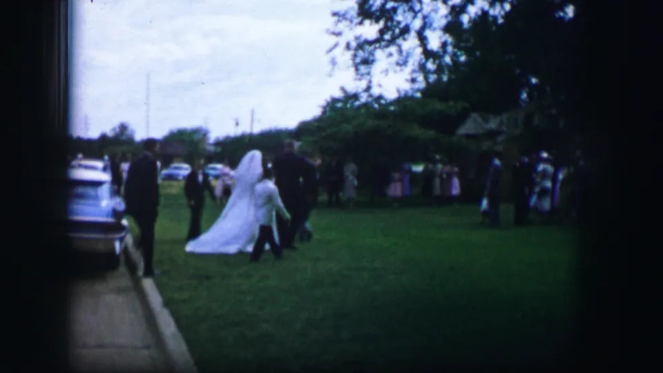 The bride crosses the grass in her white gown and veil while guests and parked cars remain visible in the Moberly wedding scene.