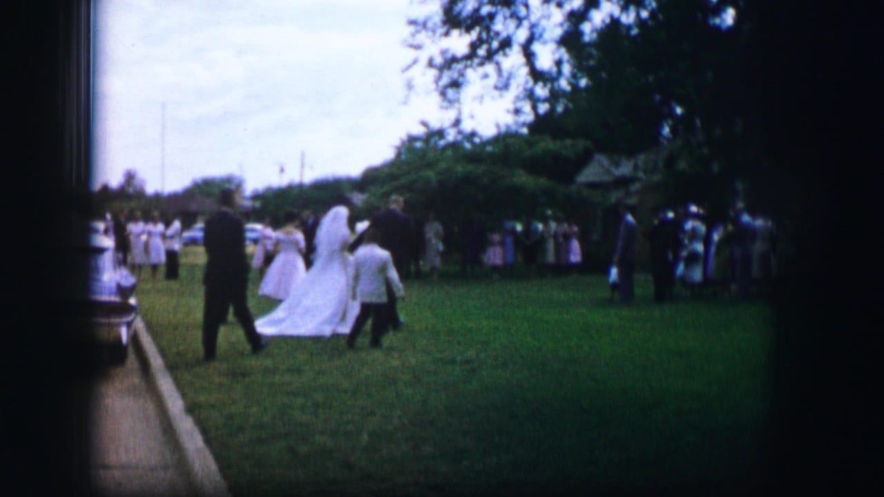 1962 Moberly, Missouri home-movie frame showing a bride in a white gown crossing a garden lawn with attendants and guests.