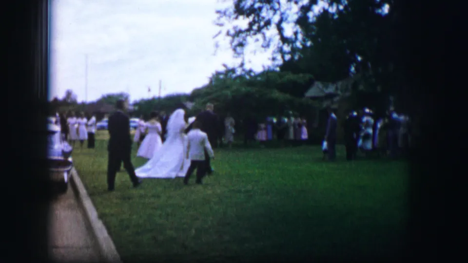 1962 Moberly, Missouri home-movie frame showing a bride in a white gown crossing a garden lawn with attendants and guests.