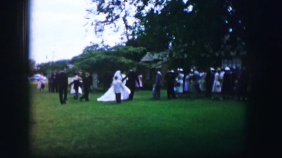 1962 Moberly home-movie frame of a bride and attendants nearing gathered guests during an outdoor garden wedding.