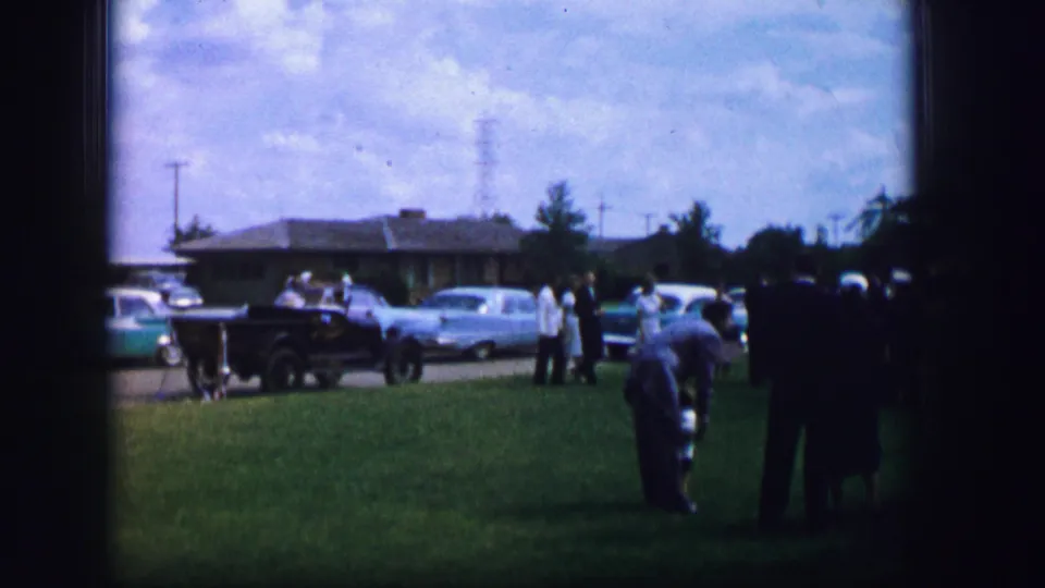 1962 Moberly, Missouri frame with wedding guests on a lawn, mid-century cars at the curb, and low houses in the background.