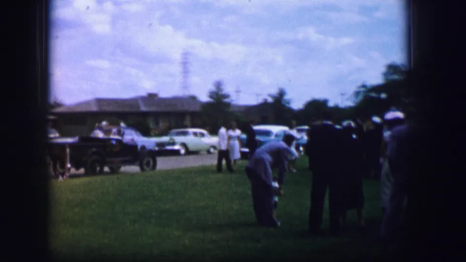 1962 Moberly, Missouri home-movie view of wedding guests on a shaded lawn, with cars and neighborhood houses beyond.