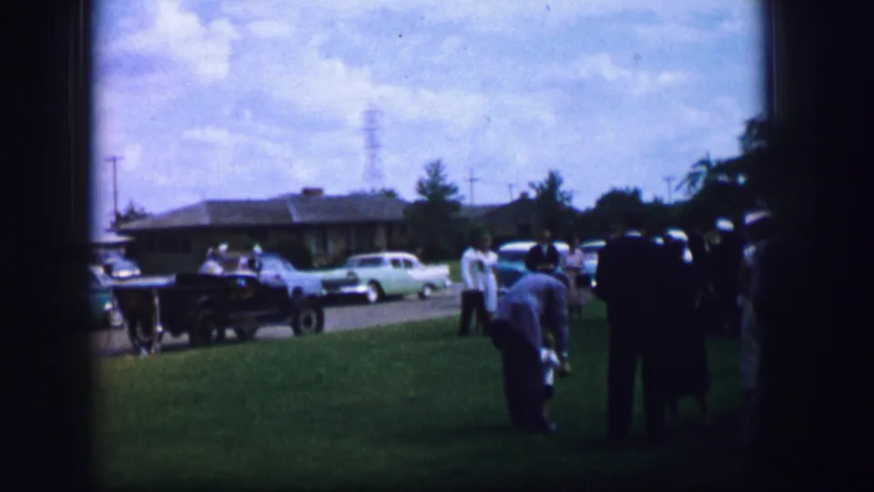1962 Moberly, Missouri frame showing guests on the lawn, one figure bending forward, with mid-century cars near the curb.