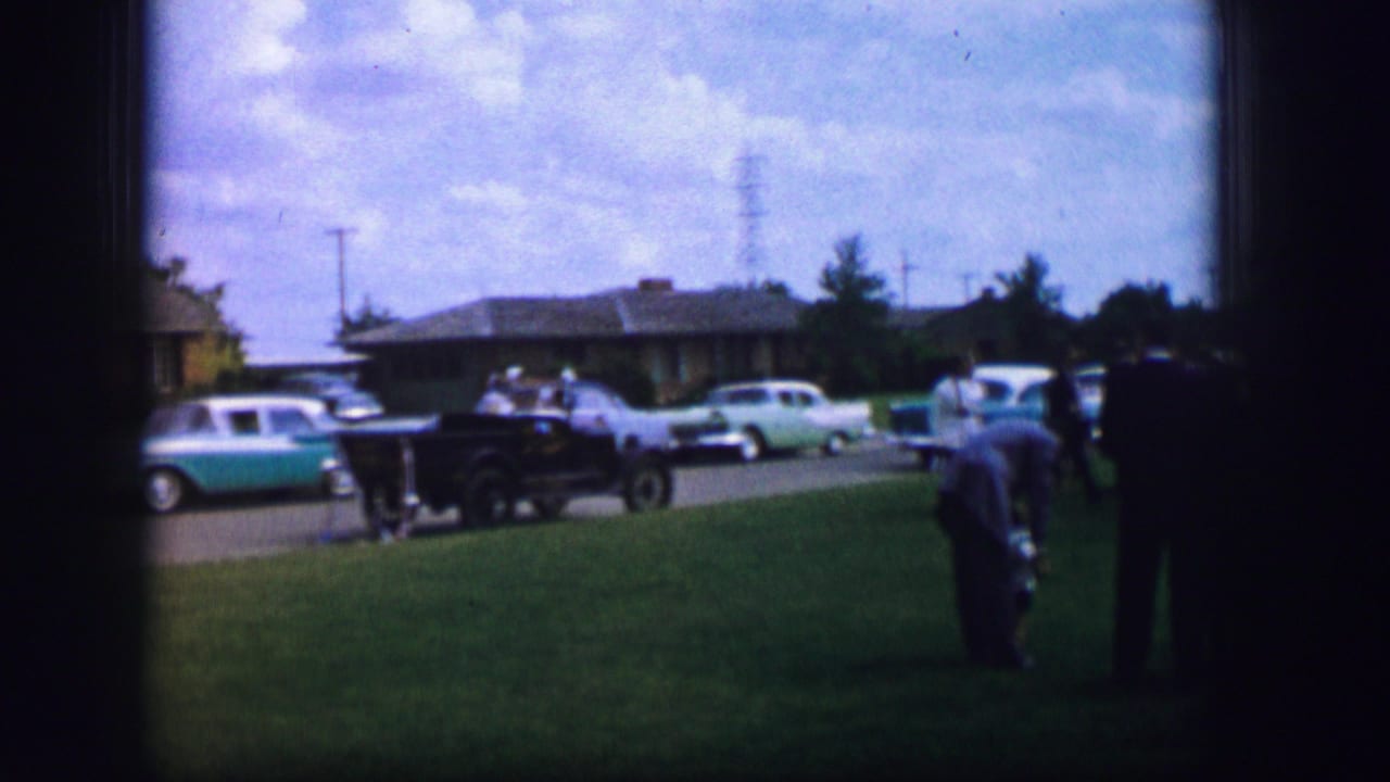 1962 Moberly, Missouri home-movie frame showing adults in a park-like lawn area with classic cars parked along the road.