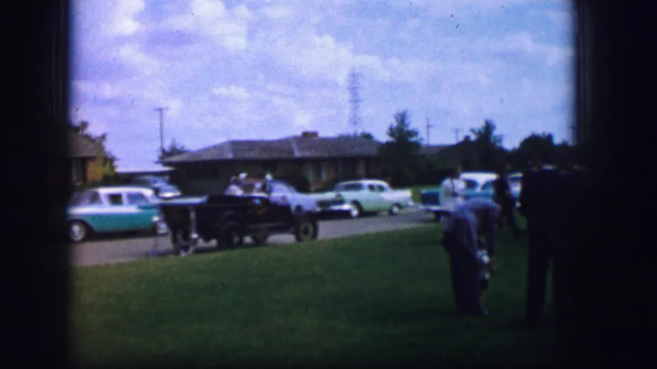 1962 Moberly, Missouri home-movie frame showing adults in a park-like lawn area with classic cars parked along the road.