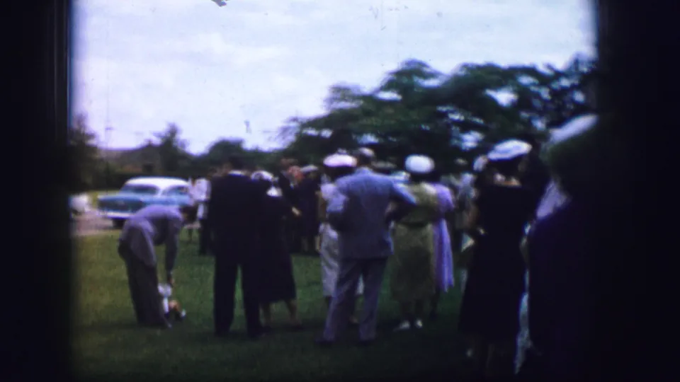 Home-movie frame from 1962 Moberly showing adults gathered in a grassy park area, some wearing hats, with parked cars behind them.