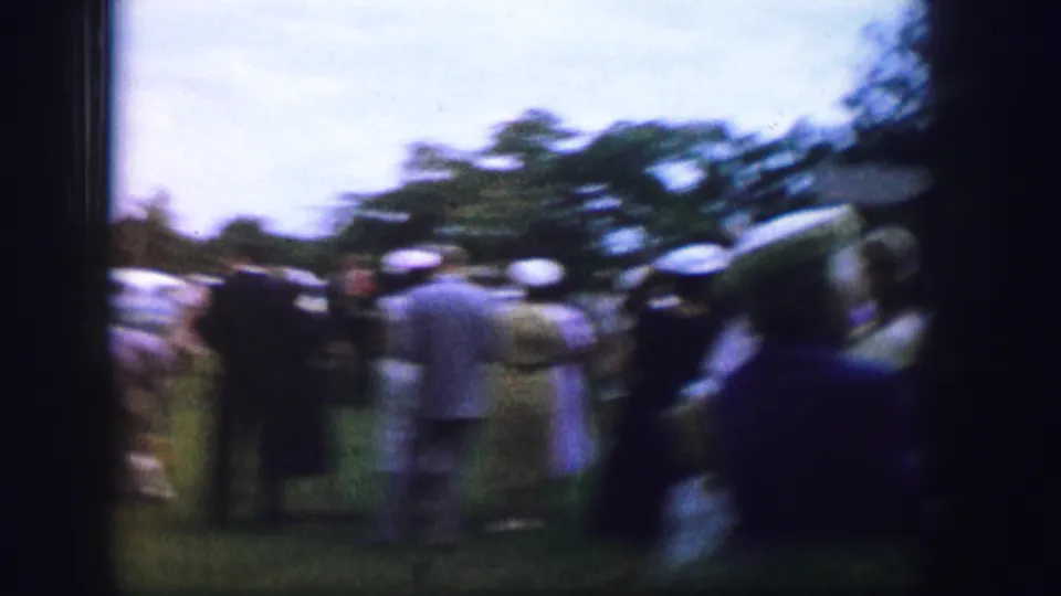 Soft, motion-blurred 8mm frame of a 1962 Moberly park gathering with adults on the lawn and classic cars in the background.