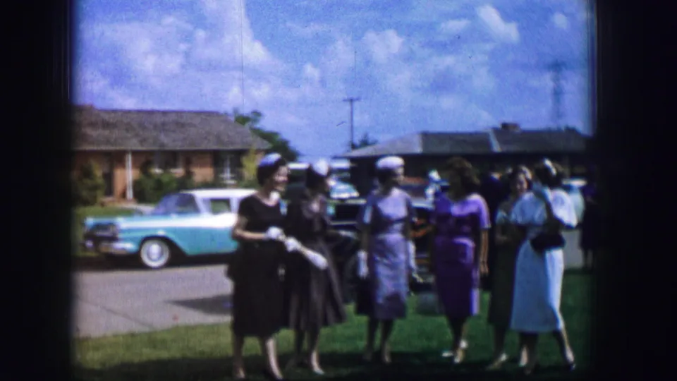 In 1962 Moberly, Missouri, women in mid-century dresses and hats gather outdoors beside parked cars and suburban houses.