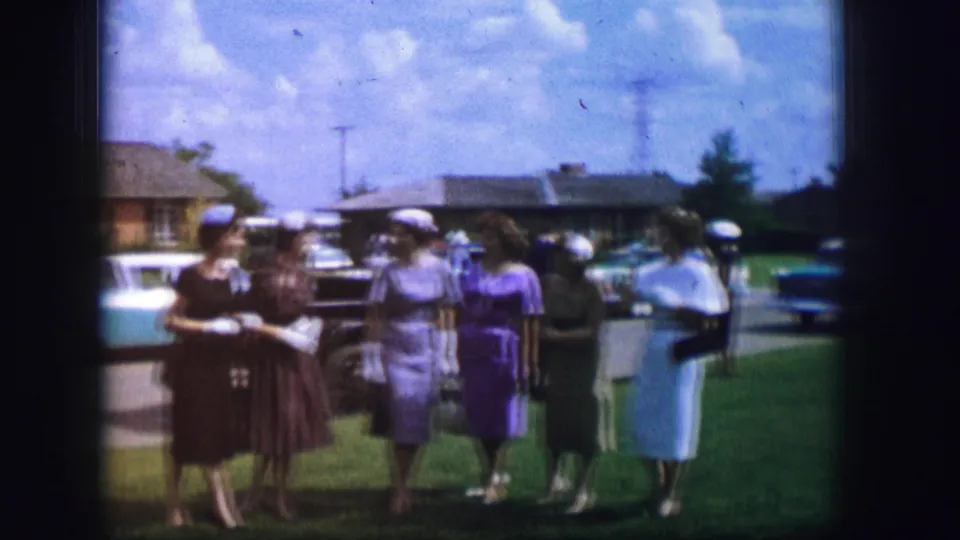 Women in 1960s dresses and hats converse while walking on a sunny lawn in the supplied Moberly, Missouri setting.