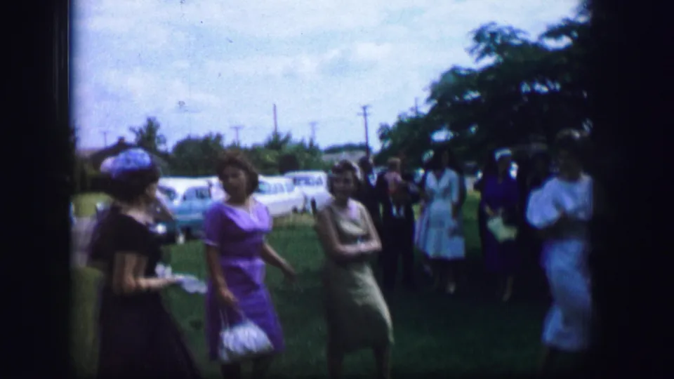 Several women in dress clothes move through an outdoor residential gathering, with cars, trees and utility poles visible behind them.