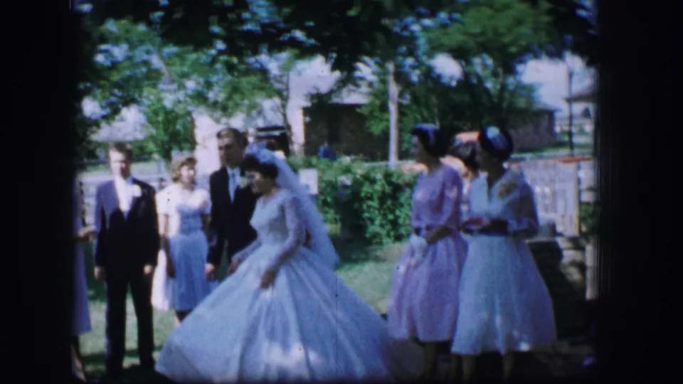 1962 Moberly, Missouri home-movie frame with bride adjusting or moving her gown as attendants stand beside her outdoors.
