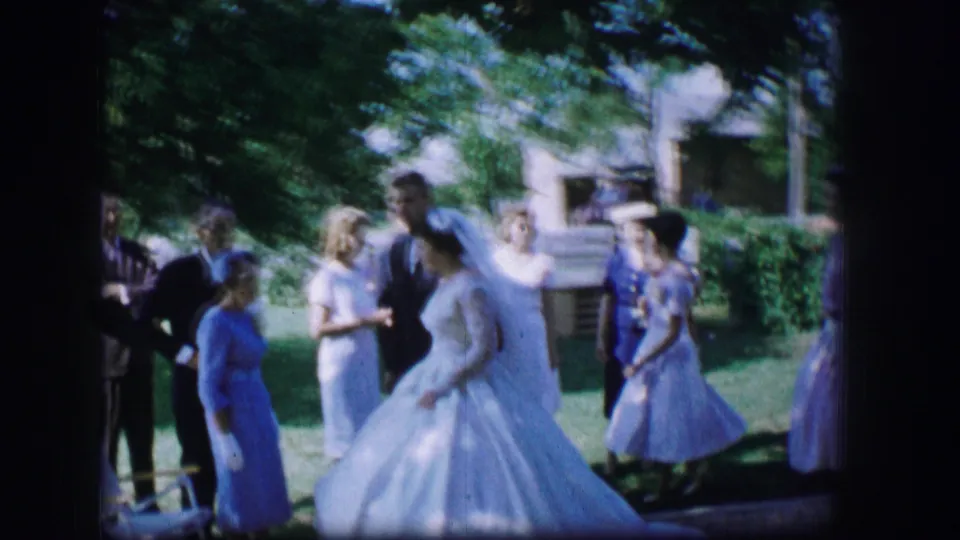 1962 Moberly wedding scene showing the bride in a full gown moving across a lawn with guests and pastel-dressed attendants behind her.
