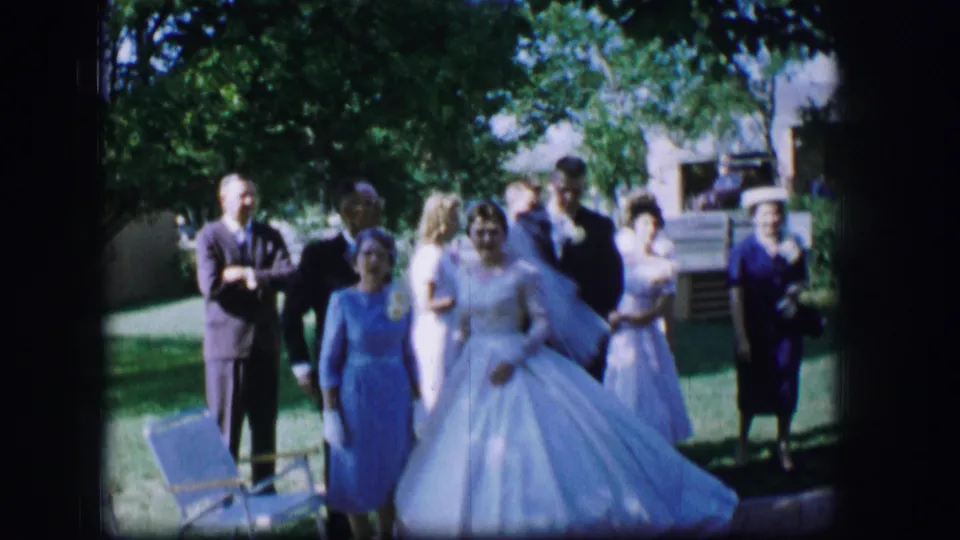 The bride reaches the groom in a garden wedding scene in Moberly, Missouri, with guests standing nearby in 1960s formal dress.