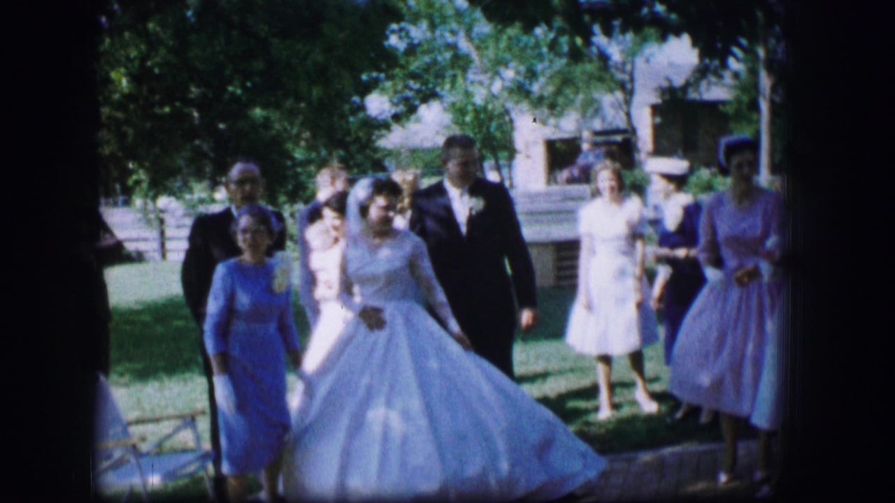 In Moberly, Missouri, 1962, a bride in a white gown and groom in a dark suit walk with guests across a green lawn.