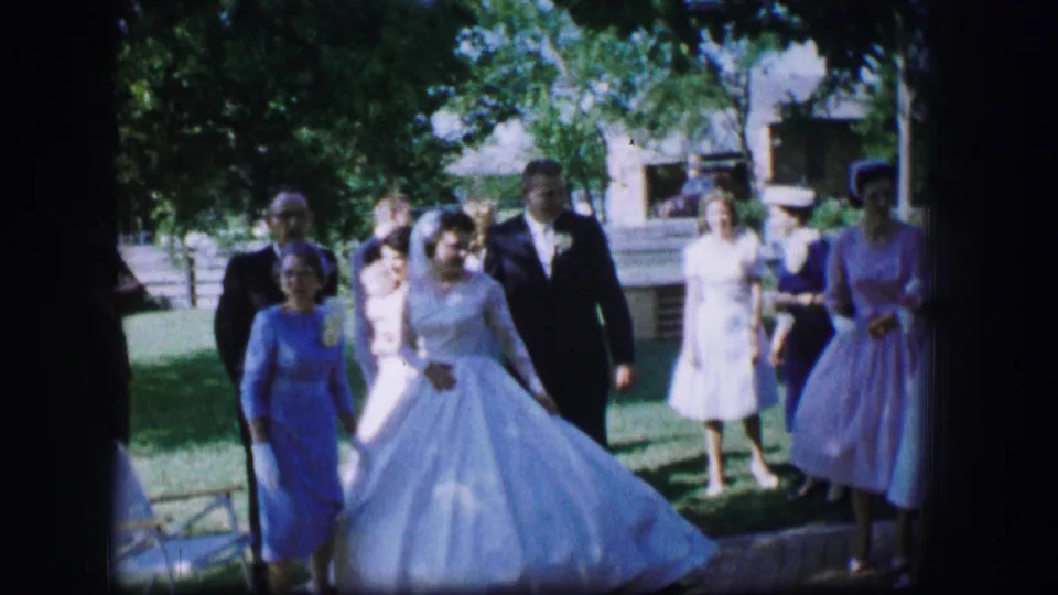 In Moberly, Missouri, 1962, a bride in a white gown and groom in a dark suit walk with guests across a green lawn.