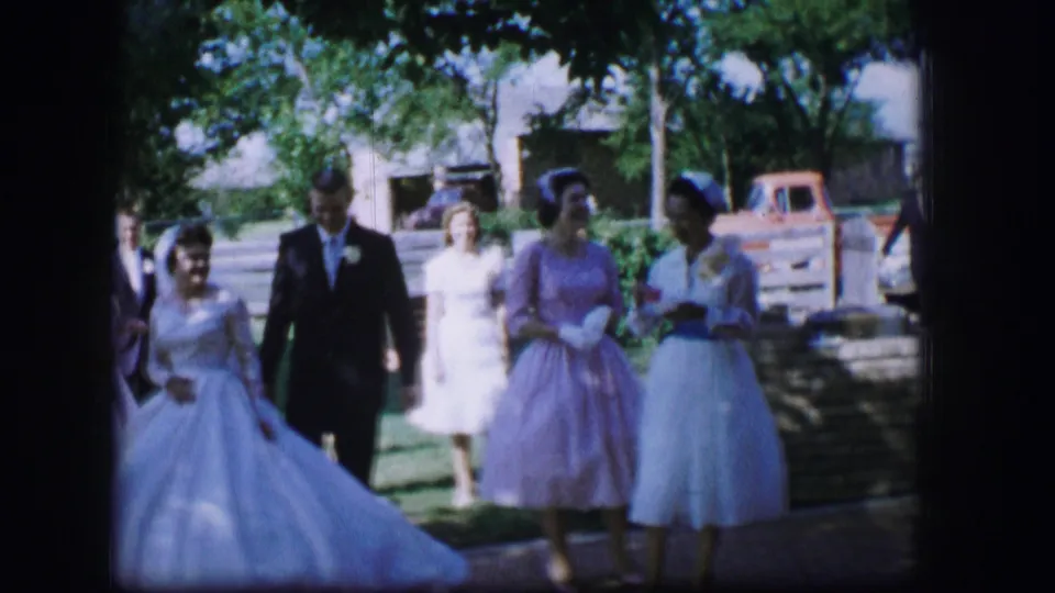The bride and groom move past guests in pastel dresses near a residential yard, trees, and a parked pickup.