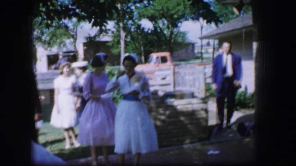 Two women in light formal dresses stand near a low stone wall while other wedding guests move through the sunny yard.