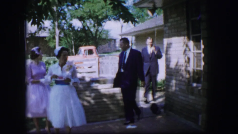 Wedding guests in formal clothing stand near a residential house, low wall, and parked pickup in the 1962 Moberly footage.